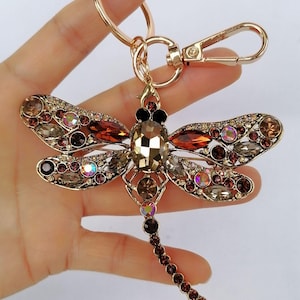 Op de afbeelding: Een goudkleurige sleutelhanger in de vorm van een libel met een karabijnhaak en sleutelring. De libel is versierd met bruine, amberkleurige en iriserende kristallen. Het lichaam van de libel is een grote, facetgeslepen, amberkleurige steen.
