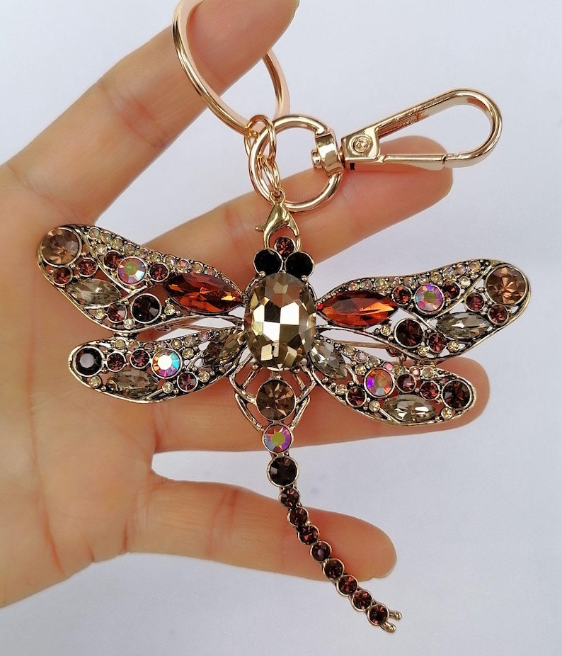 Op de afbeelding: Een goudkleurige sleutelhanger in de vorm van een libel met een karabijnhaak en sleutelring. De libel is versierd met bruine, amberkleurige en iriserende kristallen. Het lichaam van de libel is een grote, facetgeslepen, amberkleurige steen.