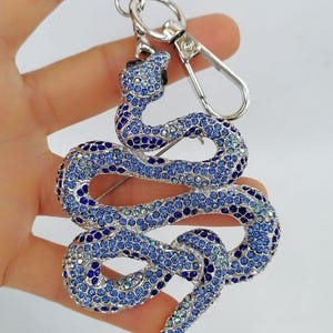 May include: A silver-toned keychain featuring a snake design encrusted with blue and clear crystals. The snake is coiled and has a detailed head and tail. The keychain includes a split ring and a lobster clasp for easy attachment.