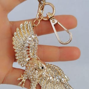 May include: Gold-toned eagle keychain, adorned with sparkling clear crystals. The eagle is depicted with outstretched wings, and a lobster clasp and key ring are attached. A decorative accessory.
