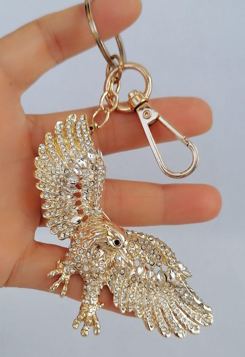 May include: Gold-toned eagle keychain, adorned with sparkling clear crystals. The eagle is depicted with outstretched wings, and a lobster clasp and key ring are attached. A decorative accessory.