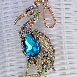 May include: A gold-toned metal keychain featuring a bejeweled bird design. The bird has a large, teardrop-shaped blue crystal and is adorned with colorful rhinestones. The keychain includes a clasp and a ring.