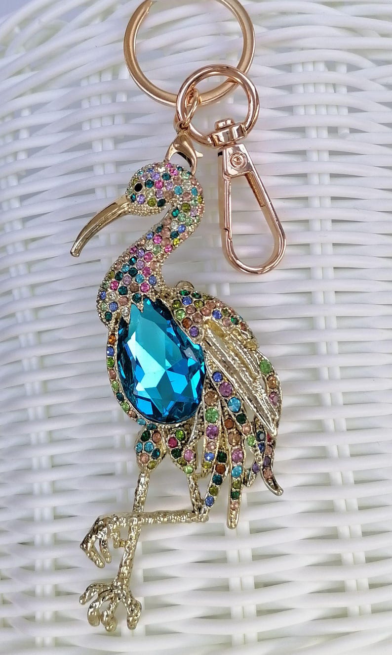 May include: A gold-toned metal keychain featuring a bejeweled bird design. The bird has a large, teardrop-shaped blue crystal and is adorned with colorful rhinestones. The keychain includes a clasp and a ring.