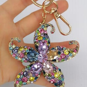 May include: A gold-toned metal keychain featuring a large, multi-colored crystal flower design. The flower is adorned with a variety of colorful crystals, including pink, blue, yellow, and purple. The keychain has a lobster clasp and a key ring.