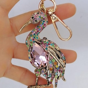 May include: A gold-toned metal keychain featuring a bejeweled bird design. The bird has a large pink gemstone on its chest and is adorned with colorful rhinestones. The keychain includes a lobster clasp and a key ring.