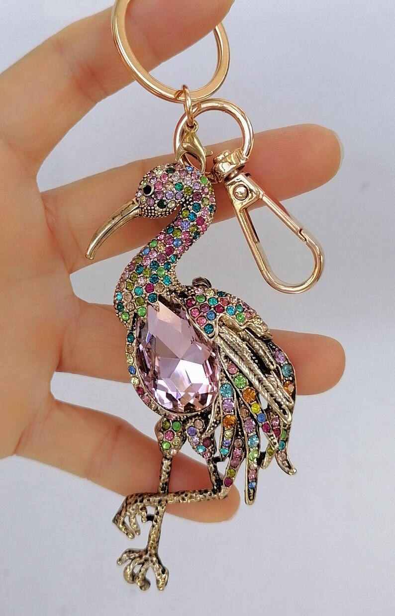 May include: A gold-toned metal keychain featuring a bejeweled bird design. The bird has a large pink gemstone on its chest and is adorned with colorful rhinestones. The keychain includes a lobster clasp and a key ring.