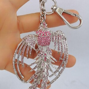 May include: A silver-toned metal keychain featuring a phoenix design. The phoenix is adorned with pink rhinestones, creating a sparkling effect. The keychain includes a lobster clasp and a key ring for easy attachment.