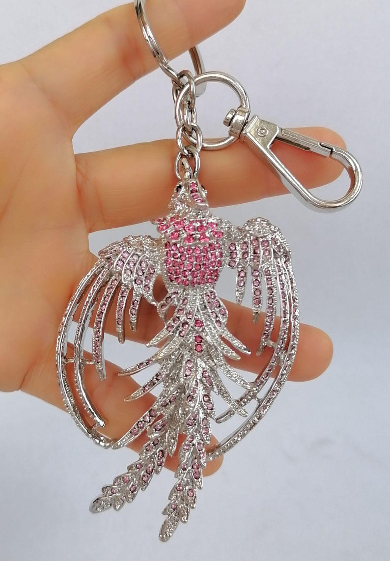 May include: A silver-toned metal keychain featuring a phoenix design. The phoenix is adorned with pink rhinestones, creating a sparkling effect. The keychain includes a lobster clasp and a key ring for easy attachment.
