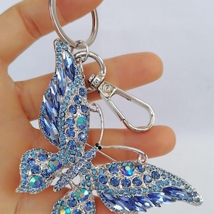 May include: A silver-toned butterfly keychain adorned with blue crystals. The butterfly's wings are detailed with various shades of blue stones, creating a sparkling effect. The keychain includes a keyring and a lobster clasp.