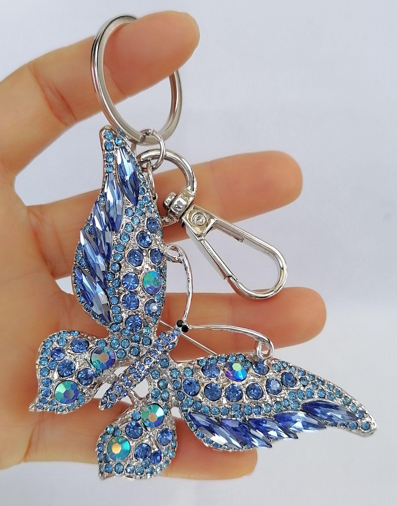 May include: A silver-toned butterfly keychain adorned with blue crystals. The butterfly's wings are detailed with various shades of blue stones, creating a sparkling effect. The keychain includes a keyring and a lobster clasp.