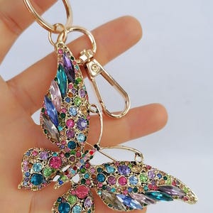 May include: A gold-colored butterfly keychain adorned with a multitude of colorful rhinestones. The butterfly's wings are embellished with various shapes and sizes of gems in shades of pink, blue, green, purple, and clear. The keychain includes a clasp and a ring.