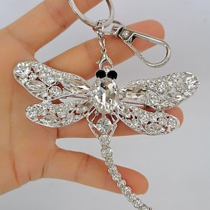Op de afbeelding: Een zilverkleurige sleutelhanger in de vorm van een libel met heldere kristallen accenten. De libel heeft gedetailleerde vleugels en een lichaam versierd met sprankelende kristallen. De sleutelhanger bevat een sluiting en een sleutelring.
