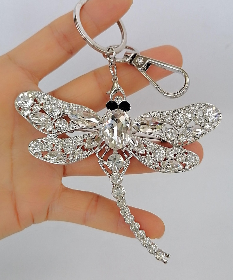 Op de afbeelding: Een zilverkleurige sleutelhanger in de vorm van een libel met heldere kristallen accenten. De libel heeft gedetailleerde vleugels en een lichaam versierd met sprankelende kristallen. De sleutelhanger bevat een sluiting en een sleutelring.