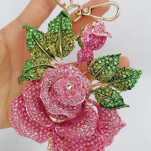 May include: A rose-shaped keychain with pink and green crystals. The rose is in full bloom, with a smaller bud and green leaves. The keychain has a gold-colored clasp and ring.