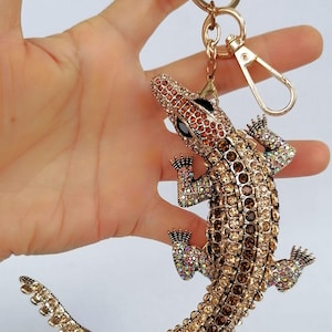 May include: A gold-toned alligator keychain, encrusted with sparkling rhinestones in shades of brown, amber, and iridescent colors. The keychain features a lobster clasp and a key ring. The alligator's eyes are black.