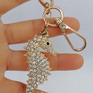 K&ouml;nnte beinhalten: Goldfarbener Seepferdchen-Schl&uuml;sselanh&auml;nger mit Karabinerverschluss. Das Seepferdchen ist mit klaren Kristallen besetzt und hat ein schwarzes Auge. Der Schl&uuml;sselanh&auml;nger wird von einem goldfarbenen Ring gehalten.