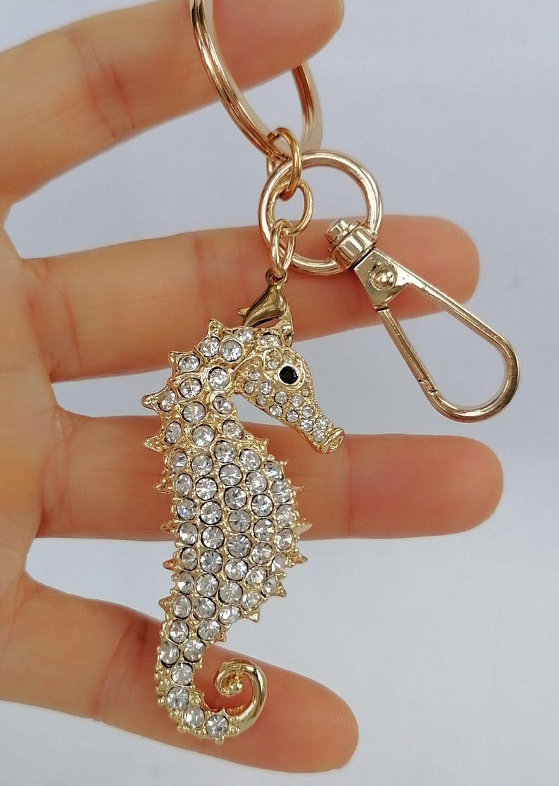K&ouml;nnte beinhalten: Goldfarbener Seepferdchen-Schl&uuml;sselanh&auml;nger mit Karabinerverschluss. Das Seepferdchen ist mit klaren Kristallen besetzt und hat ein schwarzes Auge. Der Schl&uuml;sselanh&auml;nger wird von einem goldfarbenen Ring gehalten.