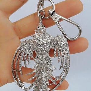 May include: A silver-colored keychain featuring a phoenix design, encrusted with numerous clear crystals. The phoenix has outstretched wings and a detailed tail, attached to a metal ring and clasp.