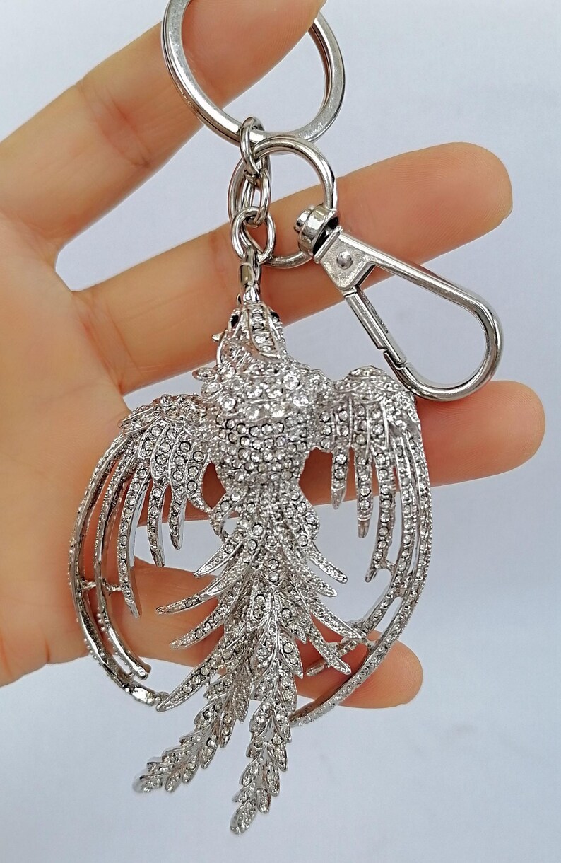 May include: A silver-colored keychain featuring a phoenix design, encrusted with numerous clear crystals. The phoenix has outstretched wings and a detailed tail, attached to a metal ring and clasp.