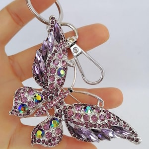 May include: A silver-toned butterfly keychain adorned with purple and iridescent rhinestones. The butterfly design features detailed wings and body, with a lobster clasp and key ring for attachment. The keychain is held in a hand.