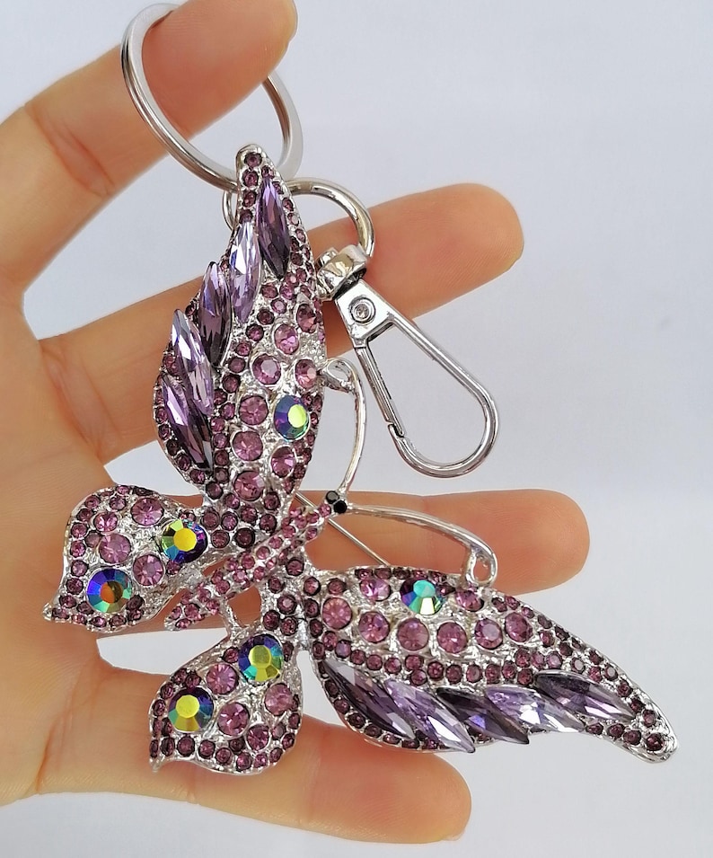 May include: A silver-toned butterfly keychain adorned with purple and iridescent rhinestones. The butterfly design features detailed wings and body, with a lobster clasp and key ring for attachment. The keychain is held in a hand.