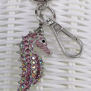 K&ouml;nnte beinhalten: Ein silberfarbener Seepferdchen-Schl&uuml;sselanh&auml;nger mit rosa und irisierenden Strasssteinen. Das Seepferdchen hat ein detailliertes Design mit einem Karabinerverschluss und einem Schl&uuml;sselring. Ein dekoratives Accessoire.