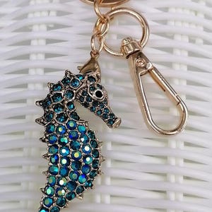 K&ouml;nnte beinhalten: Ein goldfarbener Seepferdchen-Schl&uuml;sselanh&auml;nger mit Karabinerverschluss. Das Seepferdchen ist mit blau-t&uuml;rkisen schillernden Kristallen besetzt. Der Schl&uuml;sselanh&auml;nger ist ein dekoratives Accessoire.