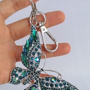 May include: A silver-toned butterfly keychain adorned with teal and green rhinestones. The butterfly's wings are detailed with various sizes of sparkling crystals. The keychain includes a clasp and a key ring.