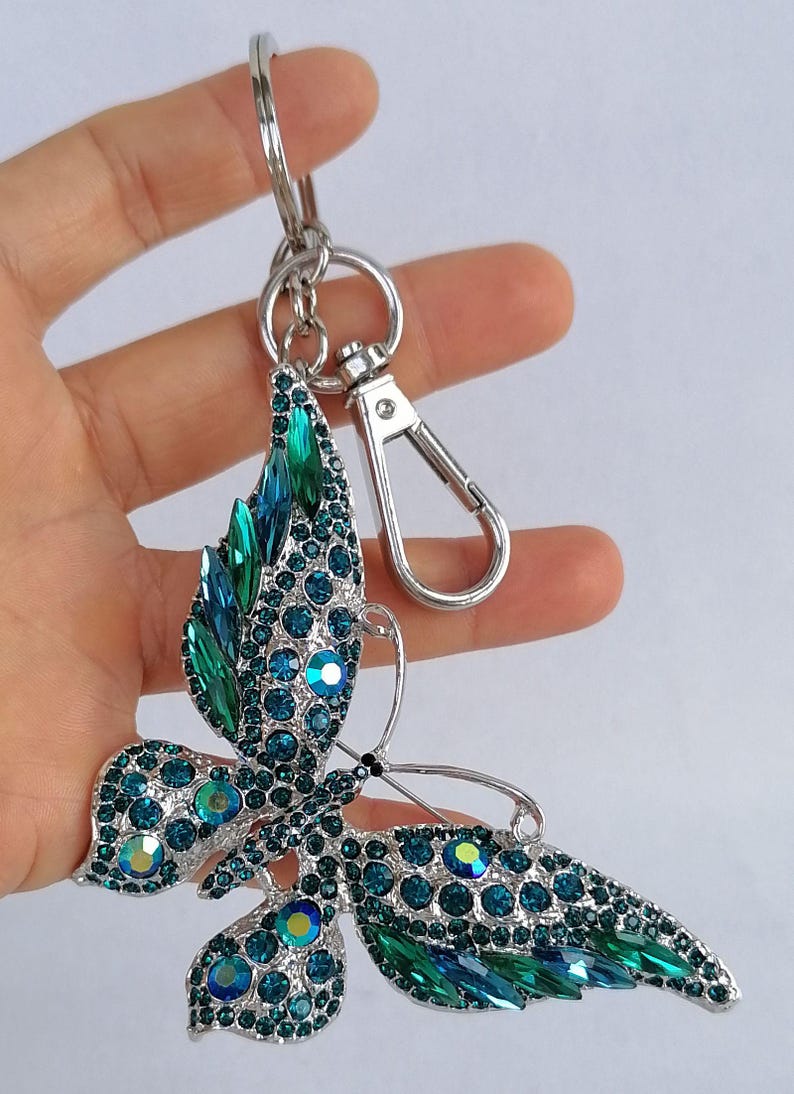 May include: A silver-toned butterfly keychain adorned with teal and green rhinestones. The butterfly's wings are detailed with various sizes of sparkling crystals. The keychain includes a clasp and a key ring.
