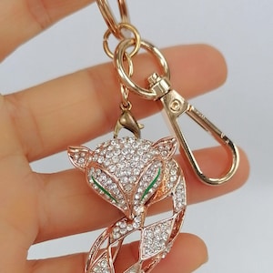May include: A gold-colored fox keychain with a lobster clasp and key ring. The fox pendant is encrusted with clear rhinestones and has green accents on the eyes. The keychain is held up by a hand.