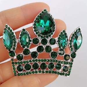May include: A silver crown brooch set with vibrant emerald green gemstones. The design includes a large central teardrop stone and smaller, round and oval gems. The brooch is held in a hand against a white backdrop.