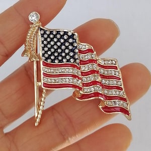 Puede incluir: Un broche dorado con la forma de la bandera estadounidense. La bandera presenta rayas rojas y blancas, un campo azul con estrellas blancas y est&aacute; adornada con pedrer&iacute;a transparente. El broche est&aacute; sujeto por un asta dorada con una parte superior decorativa.