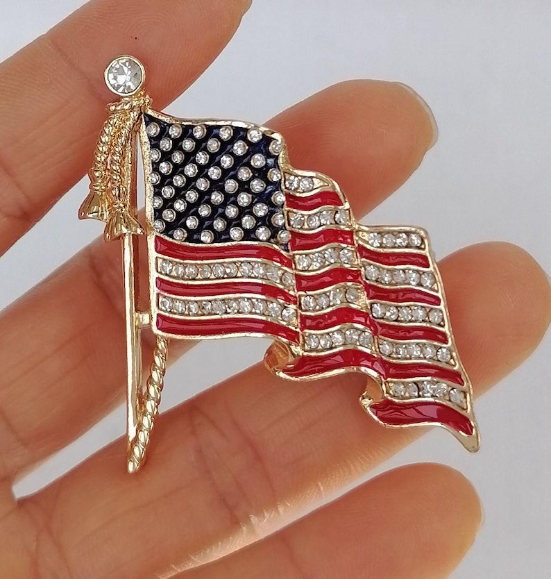 Puede incluir: Un broche dorado con la forma de la bandera estadounidense. La bandera presenta rayas rojas y blancas, un campo azul con estrellas blancas y est&aacute; adornada con pedrer&iacute;a transparente. El broche est&aacute; sujeto por un asta dorada con una parte superior decorativa.