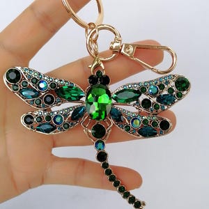 Op de afbeelding: Een goudkleurige metalen sleutelhanger in de vorm van een libel, versierd met groene en blauwgroene kristallen. De vleugels van de libel zijn versierd met ronde en druppelvormige kristallen. De sleutelhanger heeft een karabijnhaak en een sleutelring.