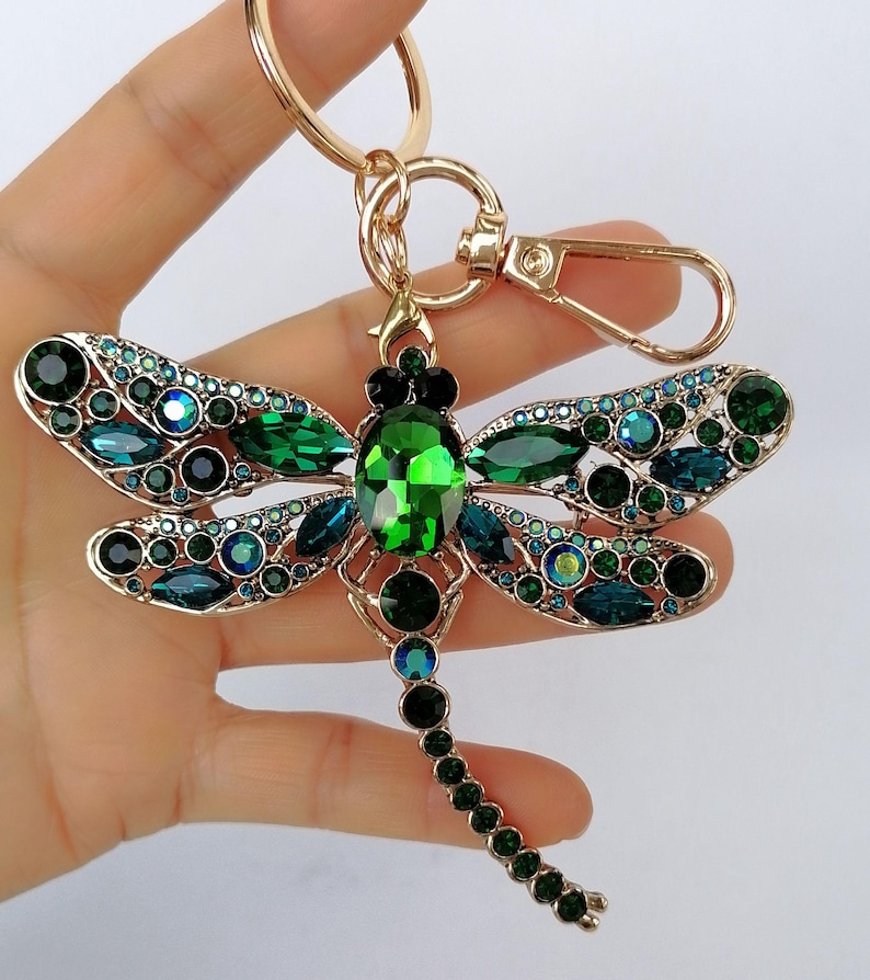 Op de afbeelding: Een goudkleurige metalen sleutelhanger in de vorm van een libel, versierd met groene en blauwgroene kristallen. De vleugels van de libel zijn versierd met ronde en druppelvormige kristallen. De sleutelhanger heeft een karabijnhaak en een sleutelring.