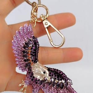 May include: A decorative eagle keychain with a gold-tone clasp and ring. The eagle is embellished with purple and black rhinestones, with a silver-colored head and gold-colored talons.