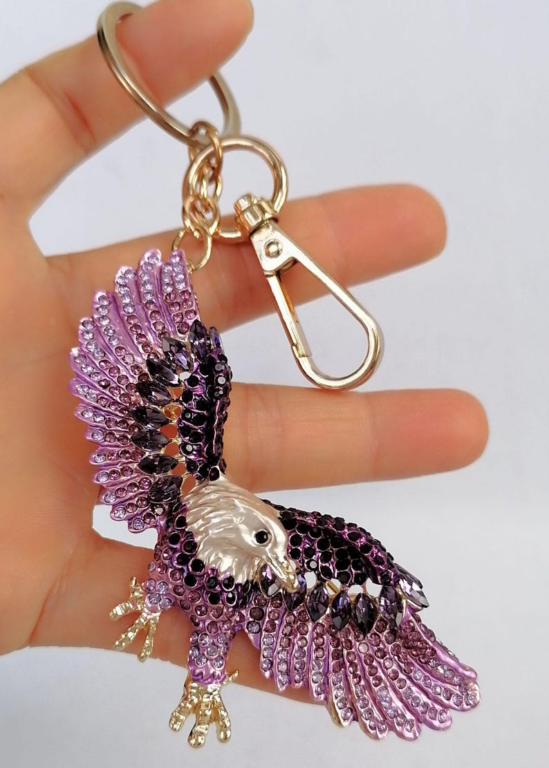 May include: A decorative eagle keychain with a gold-tone clasp and ring. The eagle is embellished with purple and black rhinestones, with a silver-colored head and gold-colored talons.
