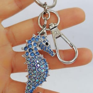 May include: A silver-toned seahorse keychain with a lobster clasp and key ring. The seahorse is encrusted with blue rhinestones. The keychain is held in a hand against a white background.
