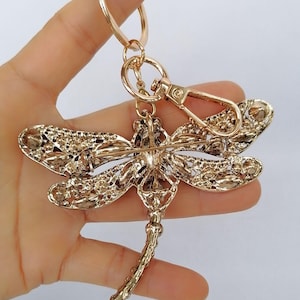 Op de afbeelding: Een goudkleurige sleutelhanger in de vorm van een libelle. De libelle heeft ingewikkelde details op de vleugels en het lichaam, met een karabijnhaak en een sleutelring. De sleutelhanger wordt tegen een lichte achtergrond gehouden.