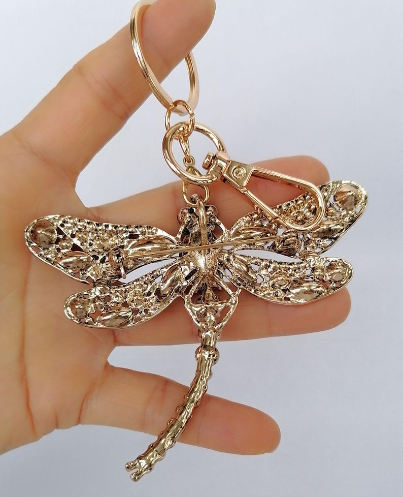 Op de afbeelding: Een goudkleurige sleutelhanger in de vorm van een libelle. De libelle heeft ingewikkelde details op de vleugels en het lichaam, met een karabijnhaak en een sleutelring. De sleutelhanger wordt tegen een lichte achtergrond gehouden.