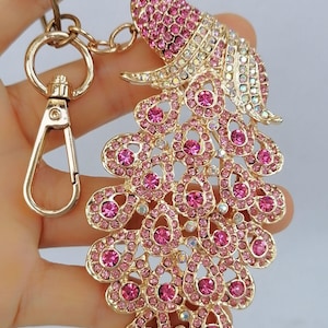 May include: Gold-toned metal peacock keychain adorned with pink and clear crystals. The peacock's tail feathers are fanned out, and the keychain includes a clasp and ring for attaching keys or accessories.