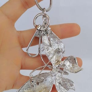 May include: Silver-toned metal keychain with a decorative floral design. The keychain features a lobster clasp, a key ring, and a detailed flower-shaped pendant with leaf accents. The pendant is attached to a pin.