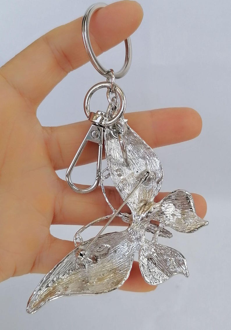 May include: Silver-toned metal keychain with a decorative floral design. The keychain features a lobster clasp, a key ring, and a detailed flower-shaped pendant with leaf accents. The pendant is attached to a pin.