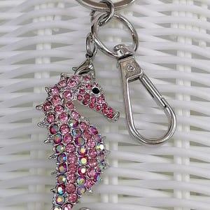 K&ouml;nnte beinhalten: Ein pinkfarbener Seepferdchen-Schl&uuml;sselanh&auml;nger mit einem silberfarbenen Metallverschluss und Schl&uuml;sselring. Das Seepferdchen ist mit rosa Strasssteinen und schillernden Akzenten besetzt. Ein dekoratives Accessoire.