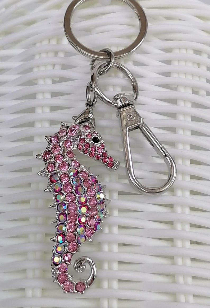 K&ouml;nnte beinhalten: Ein pinkfarbener Seepferdchen-Schl&uuml;sselanh&auml;nger mit einem silberfarbenen Metallverschluss und Schl&uuml;sselring. Das Seepferdchen ist mit rosa Strasssteinen und schillernden Akzenten besetzt. Ein dekoratives Accessoire.