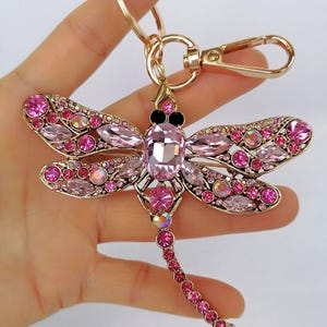 Op de afbeelding: Een goudkleurige sleutelhanger in de vorm van een libelle, versierd met roze en iriserende kristallen. De libelle heeft gedetailleerde vleugels en een lange, kralenstaart. De sleutelhanger bevat een sluiting en een ring om sleutels aan te bevestigen.