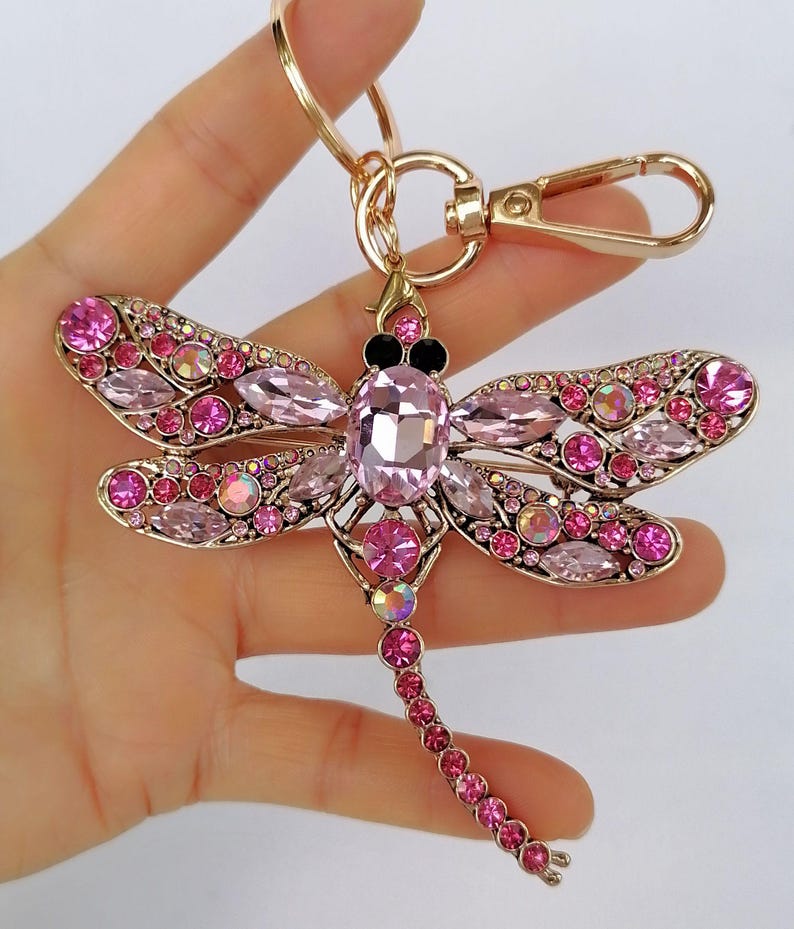 Op de afbeelding: Een goudkleurige sleutelhanger in de vorm van een libelle, versierd met roze en iriserende kristallen. De libelle heeft gedetailleerde vleugels en een lange, kralenstaart. De sleutelhanger bevat een sluiting en een ring om sleutels aan te bevestigen.