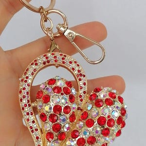 May include: Gold-toned heart-shaped keychain adorned with red and iridescent crystals. The keychain features a lobster clasp and a ring for keys. The heart design is embellished with numerous sparkling stones, creating a glamorous accessory.