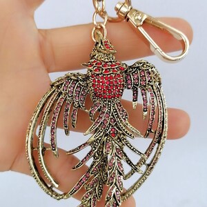 May include: A gold-toned metal keychain featuring a bird design. The bird's body is red, and its wings and tail are adorned with pink and red gemstones. The keychain includes a clasp and a ring for attaching keys.