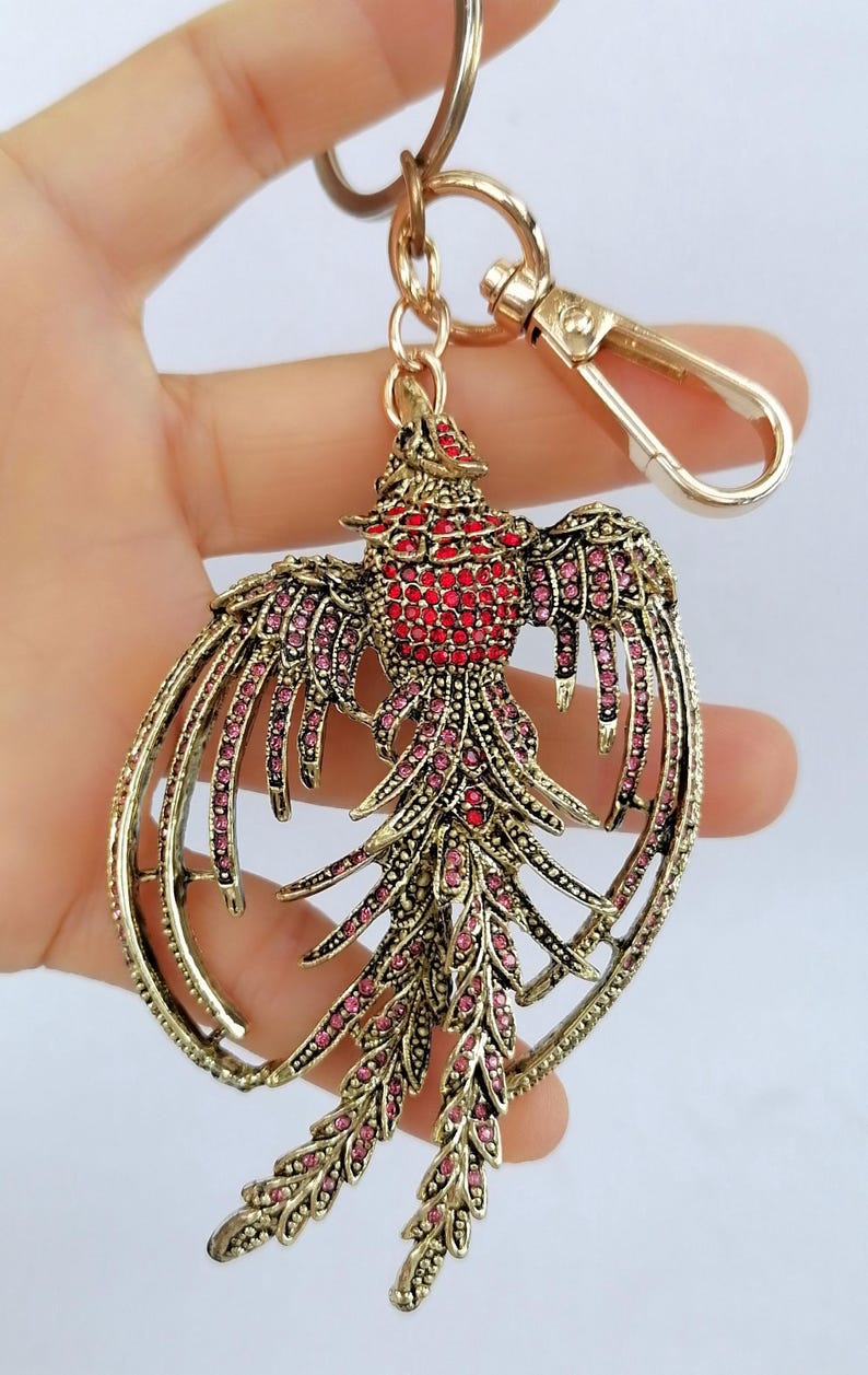 May include: A gold-toned metal keychain featuring a bird design. The bird's body is red, and its wings and tail are adorned with pink and red gemstones. The keychain includes a clasp and a ring for attaching keys.